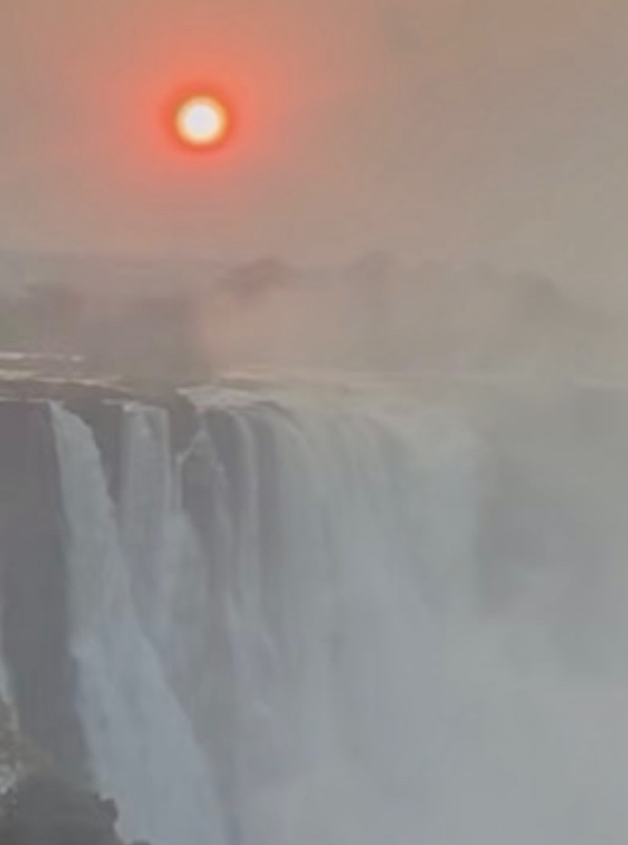 A misty view of a waterfall with the sun setting in the background, casting an orange glow.